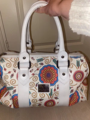 Blommig handväska från Walter Valentino - Säljer en snygg handväska från Walter Valentino med blommigt mönster i färger som röd, blå, beige och turkos. Väskan har vita handtag och detaljer, samt en metallbricka framtill. Materialet är skinnimitation och insidan är fodrad med satin. Perfekt storlek för allt du behöver.