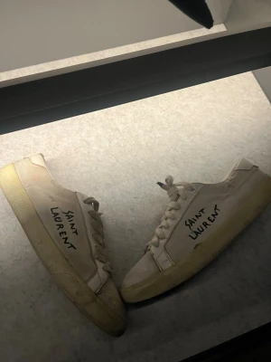 Saint Laurent vita låga sneakers - Säljer ett par vita sneakers från Saint Laurent med handskriven logga på sidan. Skorna har klassisk låg profil, vita snören och beige sula. Tillverkade i läder med stilren och minimalistisk design som passar till det mesta. Använda men har mycket kvar att ge! Skicka gärna prisförslag 
