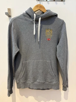 Grå hoodie från Morris Stockholm - Snygg grå hoodie från Morris Stockholm med broderad logga och brittisk flagga på bröstet. Klassisk känguruficka framtill, vita dragsnören i huvan och mjukt material som känns skönt mot huden. Perfekt för en avslappnad och stilren look.