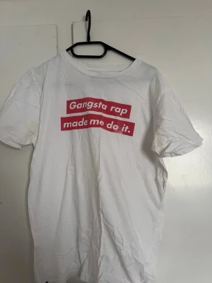 Vit t-shirt Dedicated Gangsta Rap - Vit t-shirt från Dedicated med trycket 'Gangsta rap made me do it.' i rött framtill. Tillverkad i 100% ekologisk och Fairtrade-certifierad bomull. Klassisk passform och rund hals, perfekt för en avslappnad streetwear-look.