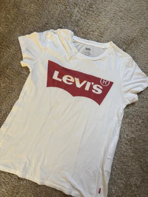 Vit Levi's t-shirt med röd logga - Säljer en klassisk vit t-shirt från Levi's med den ikoniska röda loggan tryckt framtill. T-shirten är kortärmad och är tillverkad i mjuk bomull som är skön att bära till alla dina outfits. Vid frågor hör av dig 🤍