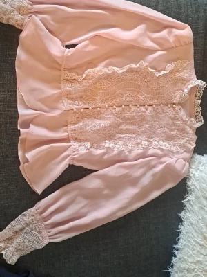 Rosa blus med spetsdetaljer - Supersöt rosa blus med romantiska spetsdetaljer framtill och vid ärmsluten. Blusen har volanger längs sidorna och långa puffärmar med breda spetsmanschetter. Perfekt för dig som gillar en feminin och vintageinspirerad stil.