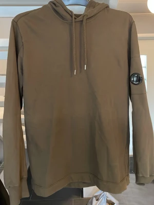 Olivgrön hoodie från C.P. Company - Snygg brun hoodie från C.P. Company med klassisk huva och dragsnören. Ärmen har den ikoniska svarta linsen med logga. Den är i väldigt bra skick tycker jag, inga defekter och syns knappt att den är använd
