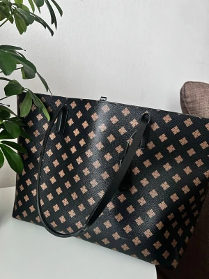 Svart shoppingväska med rutigt mönster från Malene Birger  - Stilren svart shoppingväska med beige rutigt mönster och långa svarta axelband. Väskan har rymligt ljusrosa innerfoder. Perfekt för dig som vill ha plats för allt du behöver under dagen. Den har lite slitsamma märken på såsom man ser på bilderna. Och lite märken i själva väskan men det ska nog lätt fixas man rengör den. Hade rekommenderat å ta den till en skräddare för remarna och dem eventuella märken , annars är den fortfarande snygg och hållbar. Dragkedjan går inte att stänga. 