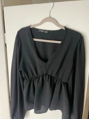 Svart v-ringad topp från Boohoo - Snygg svart blus från Boohoo med v-ringning och långa ärmar. Toppen har en markerad midja med rynk och ett lätt utställt fall. Tillverkad i ett mjukt, strukturerat tyg som ger en skön känsla och snygg siluett.