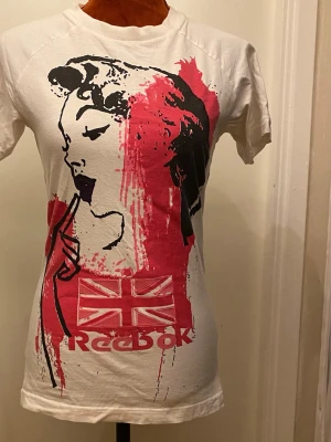 Vit Reebok t-shirt med tryck - Cool vit t-shirt från Reebok med ett stort grafiskt tryck av ett ansikte och röd färgdetalj samt brittiska flaggan. T-shirten har korta ärmar och rund hals. Perfekt för dig som gillar streetstyle och unika prints.