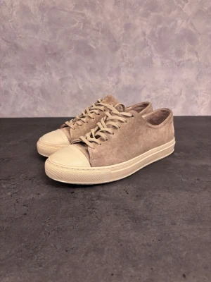 Beige mocka sneakers med gummitå - Stilrena sneakers i beige mocka med matchande snörning och vit gummisula. Skorna har en klassisk låg profil och rundad tå med förstärkt gummiparti framtill. Perfekta för dig som gillar en clean och avslappnad look.
