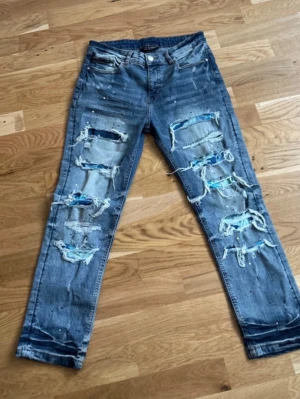 Blå slitna jeans Supply & Demand - Säljer ett par blå jeans från Supply & Demand med kraftiga slitningar och hål framtill. Jeansen har raka ben, klassisk femficksmodell och coola detaljer med trasiga partier och ljusa tvättar. Perfekta för en streetwear-look.