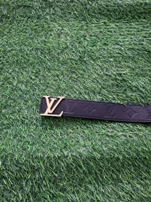 Svart Louise Vuitton skärp med L.V-spänne - Snyggt svart skärp från Louise Vuitton med klassiskt L.V-mönster präglat i lädret och ett guldigt L.V-spänne. Perfekt accessoar för att lyfta din outfit med en lyxig touch. Skärpet har en stilren och tidlös design.