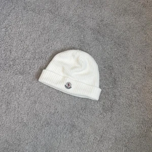 Vit ribbad mössa från Moncler - Säljer en stilren vit ribbstickad mössa från Moncler med uppvikt kant och klassisk Moncler-logga framtill. Mössan är gjord i mjukt material och har en clean look som passar soprent till alla outfits. Perfekt för dig som vill ha både värme och hype. Den är använd ett par gånger och är i bra kvalité.