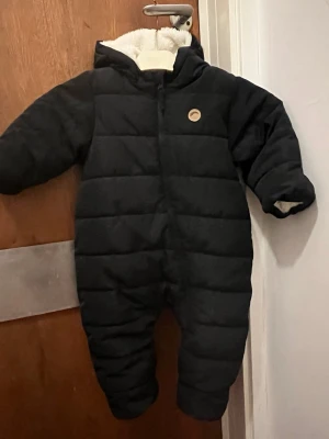 Svart fodrad täckoverall med huva - Svart quiltad täckoverall med huva och mjukt vitt foder. Dragkedja framtill och helkroppsdesign som håller värmen under kalla vinterdagar. Perfekt för att hålla sig varm ute. Enkel och stilren look.