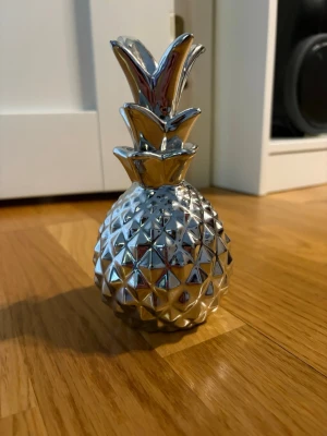 Dekorativ silverfärgad ananas - Snygg dekorationsananas i silverfärgat material med glansig yta och geometriskt mönster. Perfekt som inredningsdetalj för att ge rummet en modern touch. Tillverkad i keramik eller liknande material och har en stilren, blank finish.
