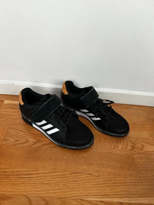Svarta Adidas träningsskor med kardborre - Säljer ett par svarta Adidas powerlift träningsskor som är lyftarskor till gymmet. Skorna har en klack för att hjälpa till vid ex knäböj. Skorna har både snörning och kardborreband för extra stöd. Ovandelen är i mesh och syntetmaterial, vilket ger bra andningsförmåga och komfort. Storlek 40 men små i storlek, passar 39! 