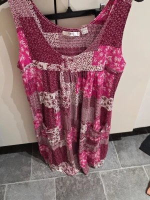 Patchworkmönstrad klänning från bpc - Mönstrad klänning i patchworkstil från bpc i nyanser av vinrött, rosa och vitt. Ärmlös modell med rund halsringning och två stora fickor framtill. Lätt och luftig, perfekt för varma dagar. Materialet känns mjukt och följsamt.