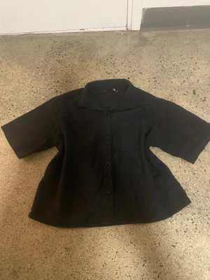 Svart kortärmad skjorta i linne - Svart skjorta med korta ärmar och avslappnad passform. Skjortan har klassisk krage, knäppning framtill och en bröstficka. Tillverkad i linne för en luftig känsla, perfekt för varmare dagar.