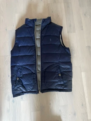 Ralph Lauren Väst 152 - Snygg vändbar dunväst från Polo Ralph Lauren med ena sidan i marinblått och andra i grått. Perfekt för våren och sommaren. Storlek 10-12/155 modellen är 157. Nypris 2299kr mitt pris 800kr. Pris går att diskuteras vid snabb affär!
