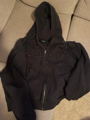 Svart zip hoodie från Axel Arigato - Säljer en svart hoodie med dragkedja från Axel Arigato. Hoodien har stor huva, broderade bokstäver på bröstet och ribbade muddar. Perfekt för en chill och stilren look. Materialet känns mjukt och skönt, och den har fickor framtill.