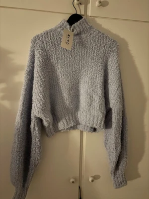 Ljusblå fluffig stickad tröja N-K-D - Supermjuk och fluffig ljusblå stickad tröja från N-K-D. Tröjan har hög hals, croppad passform och långa, lösa ärmar. Perfekt för dig som gillar en cozy och trendig look. Materialet är lurvigt och ger en riktigt mysig känsla.