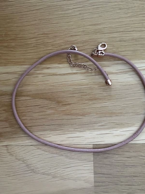 Ljusrosa armband i läderimitation - Säljer ett ljusrosa armband i läderimitation med guldfärgat spänne och kedja. Halsbandet har en enkel och stilren design som passar perfekt till olika outfits. Justerbar längd tack vare kedjan. Perfekt accessoar för att lyfta din look.