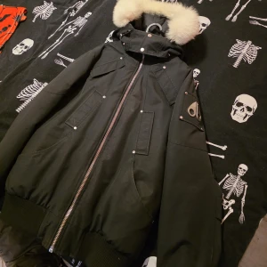 Svart dunjacka Moose Knuckles med päls - Säljer en svart dunjacka från Moose Knuckles med fluffig vit päls på huvan. Jackan har flera fickor med knappar, grov dragkedja och coolt logomärke på ärmen. Storlek M. Dragkedjan på innerfickan är trasig, har inte försökt att laga. 