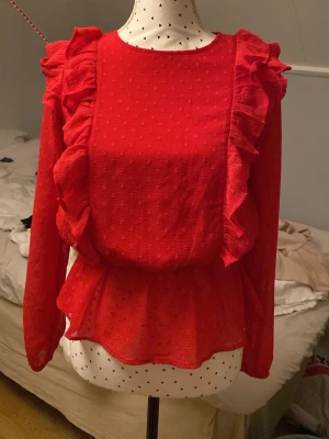 Röd volangblus med prickigt mönster - Röd blus med volanger längs axlar och framstycke. Blusen har ett diskret prickigt mönster i tyget, rund halsringning och markerad midja med lätt utsvängd nederdel. Långärmad modell i ett tunt, luftigt material.