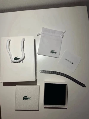 Lacoste armband  - Snyggt armband i silverfärgad metall från Lacoste med länkdesign och ingraverad krokodil på spännet. Armbandet har ett stilrent och modernt utseende, levereras med originalförpackning, påse och certifikat.
