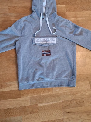 Grå hoodie från Napapijri - Snygg grå hoodie från Napapijri med stor logga och norsk flagga på bröstet. Hoodien har vit dragsko i huvan och har en stor ficka på magen. 