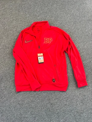 Röd Nike BP träningsjacka - Röd träningsjacka från Nike med BP-logga i gult och svart på bröstet och ryggen. Jackan har hel dragkedja, ståkrage och två sidofickor med dragkedja. Tillverkad i ett lätt och sportigt material, perfekt för träning eller uppvärmning.
