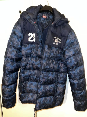 Blå camo dunjacka från Vinson - Snygg blå dunjacka från Vinson med camomönster, huva och dragkedja. Jackan har vita tryckdetaljer med siffran 23 och logga på bröstet. Perfekt för kalla vinterdagar och riktigt streetig stil.