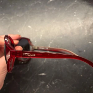 Röda solglasögon från Vogue - Stilrena solglasögon från Vogue med transparent röd båge och stora, lätt fyrkantiga glas. Snygg retrovibe och logga på sidan. Perfekta för dig som vill sticka ut med färg och form. Lätta och bekväma att bära.