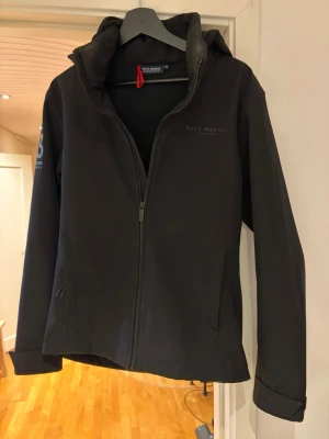 Svart vindjacka från Race Marine - Svart vindjacka från Race Marine med huva och hel dragkedja framtill. Jackan har diskret logga på bröstet och tryck med siffror och koordinater på ena ärmen. Perfekt för blåsiga dagar och enkel att matcha med olika outfits. Finns fläckar men absolut inget som förstör, går självklart att tvätta!