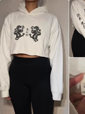 Vit cropped hoodie med draktryck från Urban Outfitters - Vit cropped hoodie med svart drakmotiv och asiatiska tecken på bröstet och ena ärmen. Hoodien har huva och långa ärmar, tillverkad i mjukt bomullsmaterial. Perfekt för en trendig och avslappnad streetstyle-look. Finns solkrämsfläckar vid kragen