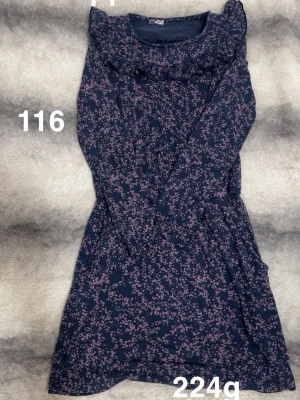 Mörkblå blommig klänning med volang - Supersöt mörkblå klänning med små blommor och volangdetalj vid halsringningen. Klänningen har långa ärmar och en mjuk, stretchig känsla. Perfekt för dig som gillar romantisk och feminin stil.🌸 Lappar borta men storlek 116 