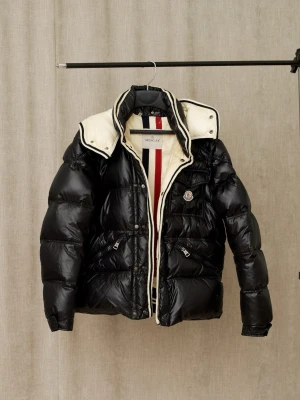 Svart dunjacka från Moncler - Snygg svart dunjacka från Moncler med glansig finish och vita detaljer på insidan. Jackan har hög krage, dragkedja och tryckknappar, samt två framfickor med dragkedja. Insidan har Monclers klassiska röd, vit och blå rand.
