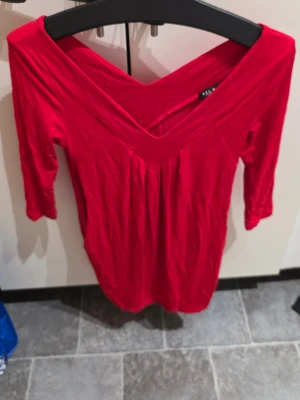 Röd v-ringad topp med trekvartsärm - Snygg röd topp med djup v-ringning och trekvartsärmar. Toppens material känns mjukt och stretchigt, perfekt för att skapa en cool och bekväm look. Passar dig som gillar färg och vill sticka ut lite extra.
