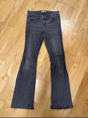 Mörkgrå bootcut jeans - Säljer ett par mörkgrå bootcut jeans med klassisk femficksdesign och lätt utsvängda ben. Jeansen har en låg midja och subtila slitningar framtill för en cool look. Perfekta för dig som gillar en avslappnad men trendig stil. Har dock ett hål i fickan.