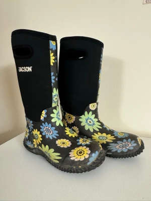 Blommiga gummistövlar i neopren från Jacson - Snygga höga gummistövlar från Jacson med svart bas och färgglatt blommönster i blått, gult och grönt. Stövlarna har praktiska handtag upptill för enkel påtagning och en robust sula som passar perfekt för regniga och kallare dagar. Materialet är en kombination av gummi och neopren. Stövlarna är mindre än 39 och uppskattas till storlek 38. 