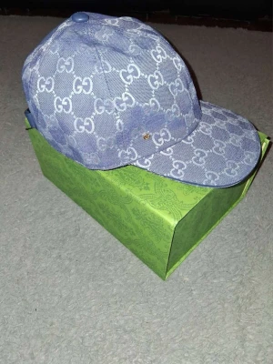 Blå Gucci keps med monogram - Säljer en blå keps från Gucci med det klassiska GG-monogrammet i vitt över hela kepsen. Kepsen har böjd skärm och är tillverkad i ett denimliknande material. Perfekt för dig som vill ha en exklusiv och trendig accessoar. Lådan får man inte vid köpet.
