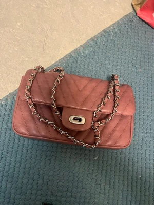 Quiltad axelväska med kedja - Säljer en snygg rosa axelväska med quiltat mönster och silverfärgad kedjerem. Väskan har ett lock med vridspänne. Perfekt för att lyfta din outfit med en trendig touch. Använd 1-2 gånger