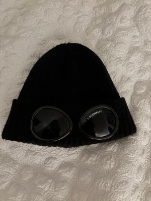 Svart C.P. Company goggle-mössa - Säljer en svart ribbstickad mössa från C.P. Company med de ikoniska goggle-linserna framtill. Mössan har uppvikt kant och är tillverkad i ett mjukt, värmande material. Perfekt för dig som vill sticka ut med en casual-inspirerad look. Mössan är äkta köpt från CPC hemsidan. Ögat på vänster har en spricka, därav det billiga priset. Man märker knappt av den. Annars perfekt skick. 