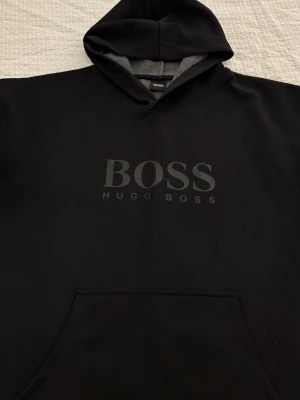 Svart Hugo Boss hoodie  - Hugo Boss hoodie i svart färg.  Stilren huvtröja från Hugo Boss med logotyp framtill. Klassisk modell med luva, kangaroo-ficka samt ribbade muddar vid ärmslut och nederkant. Stilren och nedtonad design.  • Färg: Svart   • Storlek: XL (se bild på etikett)   • Passform: Normal   • Detaljer: Luva, kangaroo-ficka, ribbade muddar   • Material: Mjuk och följsam kvalitet    Skick: Mycket bra skick – endast använd några få gånger. Inga fläckar eller hål.