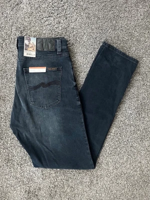 Mörkblå jeans från Nudie Jeans - Säljer ett par mörkblå jeans från Nudie Jeans med klassisk femficksdesign och svarta sömmar på bakfickan. Jeansen har en smal passform och är tillverkade i stretchigt denimtyg för extra komfort. Perfekta för dig som gillar stilrena och tidlösa jeans.