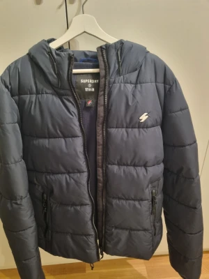 Mörkblå pufferjacka från Superdry - Snygg mörkblå pufferjacka från Superdry med huva och dragkedja framtill. Jackan har två fickor med dragkedja och ett diskret logomärke på bröstet. 