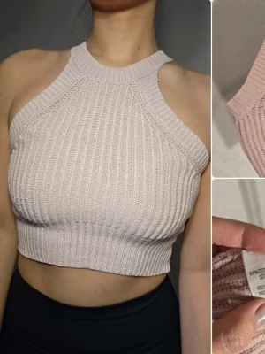 Ljusbeige ribbad croppad topp från Brandy Melville - Trendig ljusbeige croppad topp i ribbad stickad bomullsblandning. Hög halsringning och ärmlös design med breda axelband ger en sportig och modern look. Perfekt att styla med höga jeans eller kjol. Tråd vid insidan har gått upp