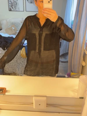 Svart blus från Second Female Copenhagen – storlek M - Stilren blus från Second Female Copenhagen i svart, storlek M. Lätt transparent med lång ärm. Endast använd ett fåtal gånger och i mycket bra skick.
