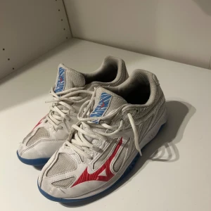 Vita Mizuno inomhusskor med röd logga - Säljer ett par vita Mizuno inomhusskor med röd logga på sidan och blå detaljer på plösen och sulan. Skorna har meshpaneler för bra ventilation och en slitstark sula som passar perfekt för sporthallar. Snygg och sportig design som sticker ut. Använda ett tag men funkar precis som vanligt. 