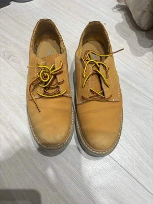 Beige loafers från Timberland - Säljer ett par beige loafers från Timberland i nubuckläder. Skorna har kontrasterande gula och bruna snören samt en ljus, robust sula. Timberland-loggan är präglad på sidan. Perfekta för dig som gillar klassisk och avslappnad stil.