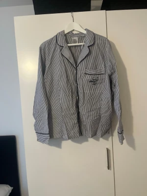 Randig pyjamaströja med ficka - Klassisk långärmad pyjamaströja med smala blå och vita ränder. Skjortmodell med krage, knappstängning och bröstficka med broderad detalj. Svarta passpoaler längs krage och ficka. Perfekt för chilliga kvällar hemma.