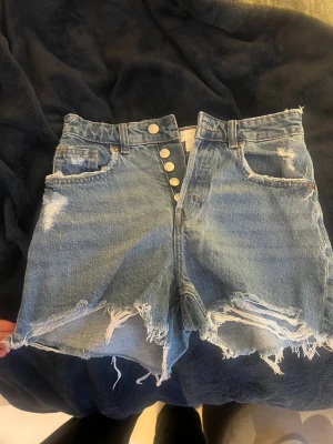 Högmidjade blå jeansshorts med knappar - Snygga högmidjade jeansshorts i blå denim med slitna detaljer och fransig kant. 