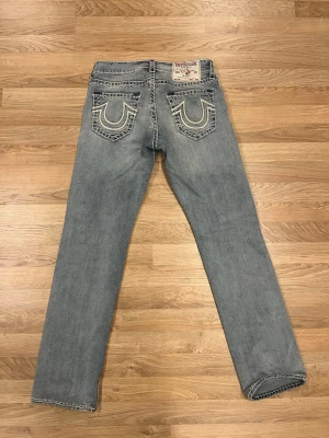True Religion jeans - Säljer ett par klassiska blå jeans från True Religion med raka ben och tydliga kontrastsömmar. Jeansen har ikoniska bakfickor med broderad logga och patch i midjan. Finns liten fläck (se bild) annars i ny skick  storlek 31
